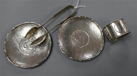 Two white metal dishes each inset with Chinese coin, a pair of Chinese sugar tongs and a napkin ring.